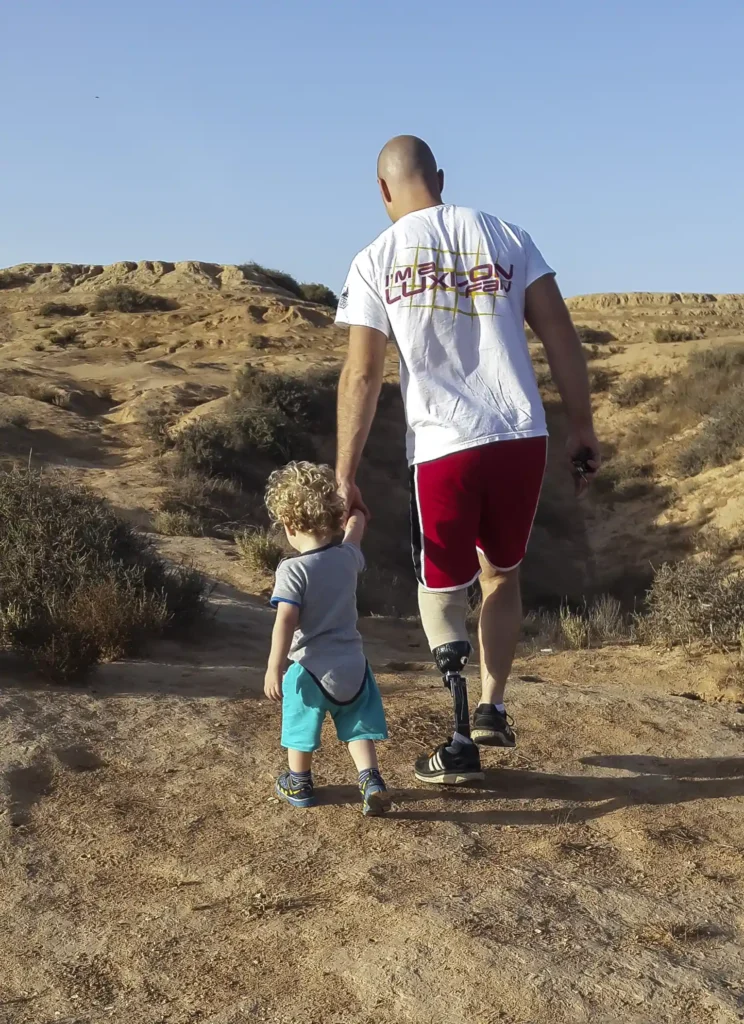 An adult with a prosthetic leg and a young child hold hands as they walk together on a rugged, desert-like trail under a clear blue sky. The adult is wearing a white T-shirt, red shorts, and black athletic shoes, while the child is dressed in a gray T-shirt, turquoise shorts, and sandals. They are walking away from the camera, creating a sense of connection and adventure against the natural landscape