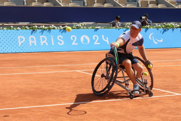Wheelchair tennis player prepares to hit a ball during a match on a clay court. The athlete, wearing a cap and sports attire, holds a racket in one hand while gripping two additional balls in the other. In the background, a blue banner with 'PARIS 2024' and the Paralympic symbol is visible, symbolizing the Paris 2024 Paralympic Games. Spectators and photographers can be seen in the stadium seats. Photo Credit - Keren Isaacson
