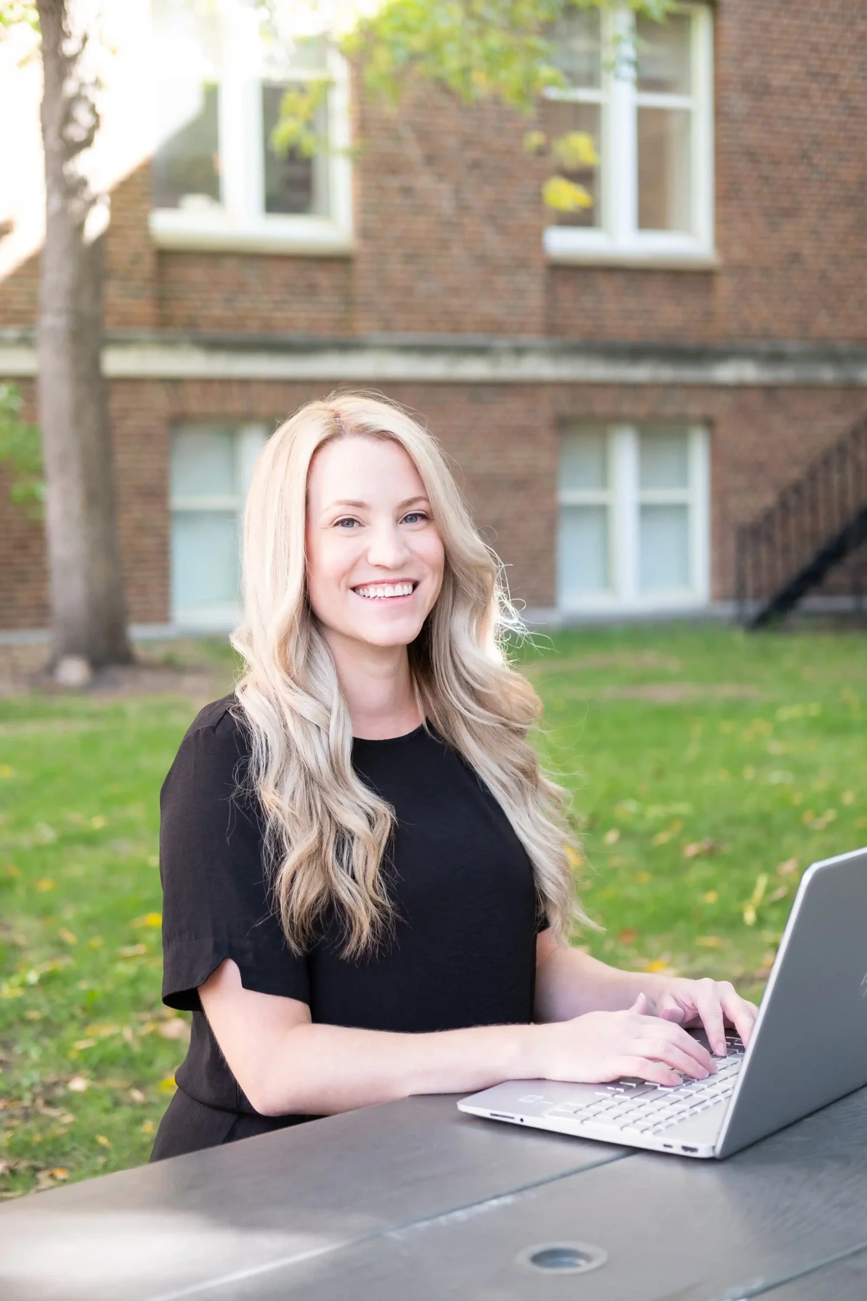 The image shows a woman sitting outdoors at a table with a laptop. She has long, wavy blonde hair and is wearing a black short-sleeved top. The setting is likely a park or a garden with a grassy area, trees, and a brick building with windows in the background. The woman is smiling warmly at the camera, giving a friendly and approachable expression. She appears to be working on her laptop. The overall mood of the image is professional yet relaxed.