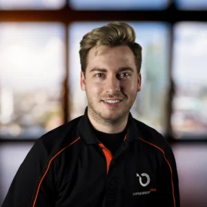 The image shows a person smiling at the camera, wearing a black polo shirt with orange accents along the shoulders and collar. The shirt features a logo on the left side that reads "companion plus" with a stylized circular symbol. The person has light skin, short blonde hair styled upward, and a light beard. The background is slightly blurred, showing large windows with a soft, cityscape view.