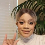 A woman is smiling at the camera, making the American Sign Language gesture for "I love you" with her hand. She has short, two-toned hair with darker roots and lighter ends, styled in a sleek middle part. She is wearing hoop earrings and a white, high-necked top. In the background, there is a window with white blinds and a large, leafy green plant. The lighting is soft and warm.