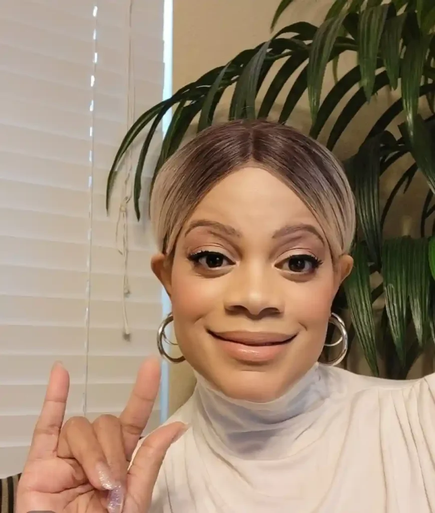 A woman is smiling at the camera, making the American Sign Language gesture for "I love you" with her hand. She has short, two-toned hair with darker roots and lighter ends, styled in a sleek middle part. She is wearing hoop earrings and a white, high-necked top. In the background, there is a window with white blinds and a large, leafy green plant. The lighting is soft and warm.