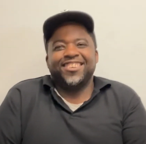 Smiling man wearing a black cap and dark shirt against a plain background