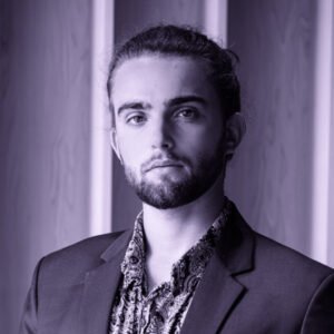 The image appears to be a black-and-white or grayscale portrait of a man with a beard and long hair tied back, wearing a suit jacket over a patterned shirt. He has a calm, neutral expression and is standing against a backdrop of vertical wooden panels. The lighting highlights his facial features, giving the image a professional yet artistic vibe.