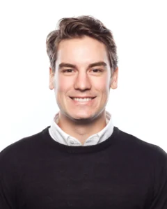 The image shows a smiling young man with short, light brown hair styled neatly. He is wearing a black crew-neck sweater over a white collared shirt. The background is completely white, giving the image a clean and professional look.