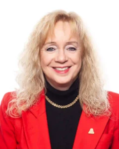 The image shows a woman with blonde, shoulder-length curly hair. She is smiling and wearing a red blazer over a black turtleneck. She has a gold chain necklace and a small pin on the left side of her blazer. The background is plain white.