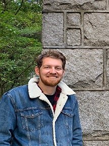 The image shows a man standing in front of a stone structure, possibly outdoors. He has light brown hair, a beard, and is smiling. He is wearing a denim jacket with a white fleece collar and a plaid shirt underneath. The background includes trees and greenery, suggesting a natural setting.