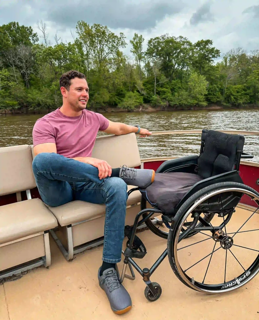 A man in a pink t-shirt and jeans is seated on a boat next to a wheelchair. He is smiling and appears relaxed, with one leg crossed over the other. The background shows a calm river surrounded by lush, green trees under a cloudy sky. The wheelchair is placed next to him, indicating that he uses it for mobility, but he is currently sitting on the boat bench. The scene conveys a peaceful, outdoor experience.