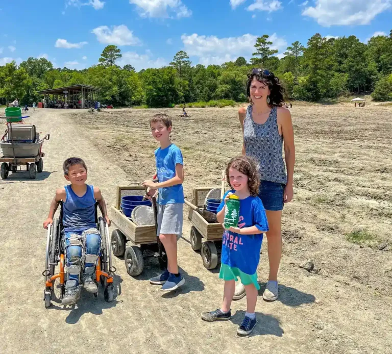 The image shows a group of three children and an adult standing outdoors on a sunny day. One child is in a wheelchair, smiling and wearing a blue tank top, while the other two children, one boy and one girl, are standing nearby, each holding onto a wooden cart filled with buckets and tools. The adult is smiling and standing next to the children, wearing a sleeveless patterned top and shorts. The setting appears to be a farm or an open field, with trees in the background and a few other people visible in the distance. The sky is bright blue with scattered clouds.