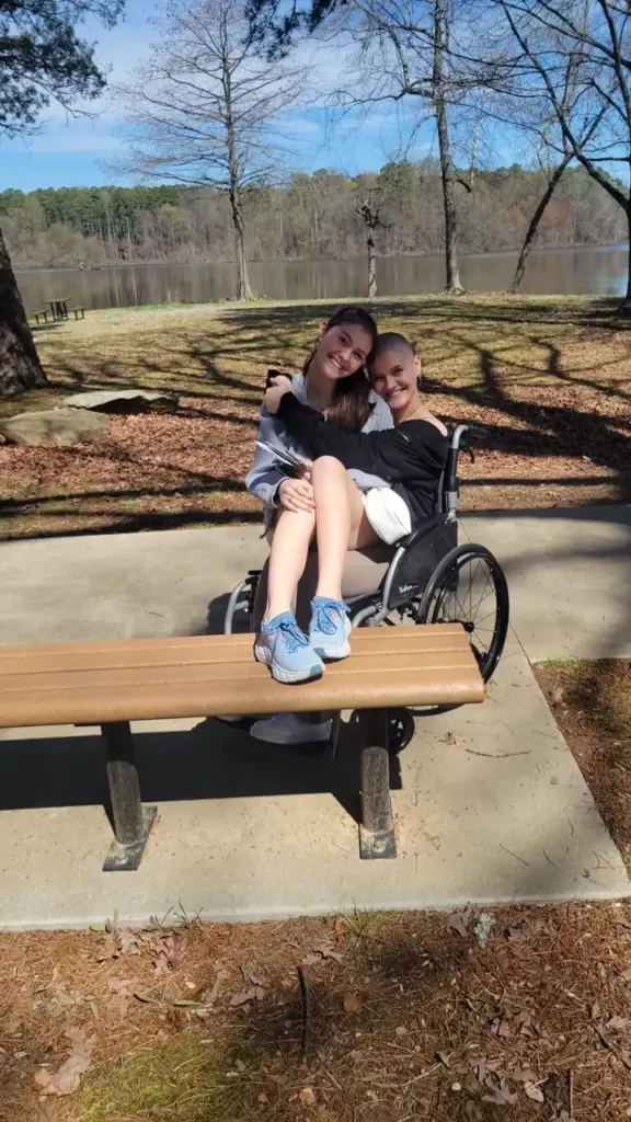 The image shows two people, one sitting on a park bench and the other in a wheelchair beside them. The person in the wheelchair is leaning towards the one on the bench, and they both have their arms around each other in an affectionate embrace. The person sitting on the bench has one leg draped over the other's lap. It appears to be a sunny day in a peaceful outdoor setting by a lake, with trees in the background, some of which are bare. The overall mood is warm and friendly, reflecting a sense of closeness between the two individuals.