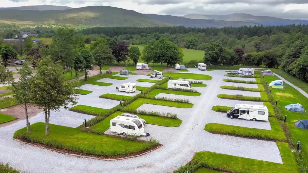 This image shows a scenic holiday park designed for caravans and camper vans, set in a lush, green environment with rolling hills and distant mountains in the background. The park has neat, gravelled parking spaces for caravans, separated by trimmed grass and hedges, creating a tidy and organized look. Surrounding the park, there are patches of trees and greenery, and the scene has a peaceful, countryside atmosphere, with a cloudy sky adding to the serene mood. Several caravans and motorhomes are parked, with some tents set up nearby.
