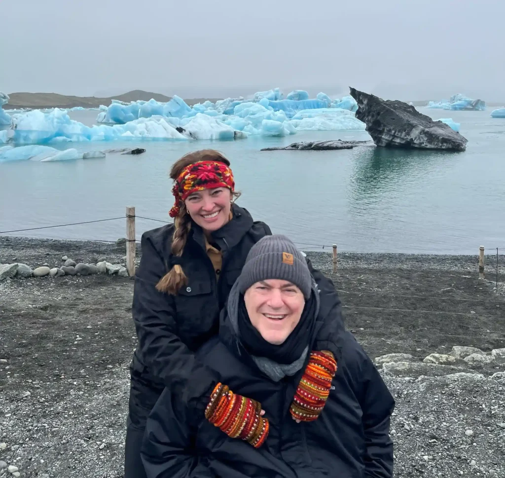 This image depicts a smiling couple bundled up in warm clothing, standing in front of an incredible backdrop of blue icebergs floating on a calm body of water. The scene looks chilly and serene, with the cloudy sky blending with the icy surroundings, likely set in a location known for its glaciers or icebergs. The individuals are dressed in black jackets, with colorful knit accessories like hats, headbands, and mittens. The overall mood is happy and adventurous, as they appear to be enjoying their time in a stunning, cold, natural landscape.