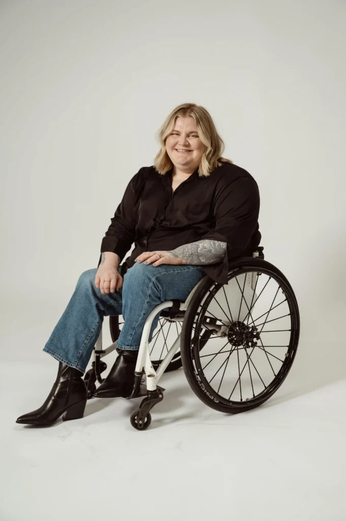 A person with blonde shoulder-length hair smiles while seated in a wheelchair. They are wearing a black long-sleeve blouse, blue jeans, and black ankle boots with a slight heel. Their left arm has visible tattoos, and they are resting their hands on their lap. The background is a neutral, light-colored backdrop.