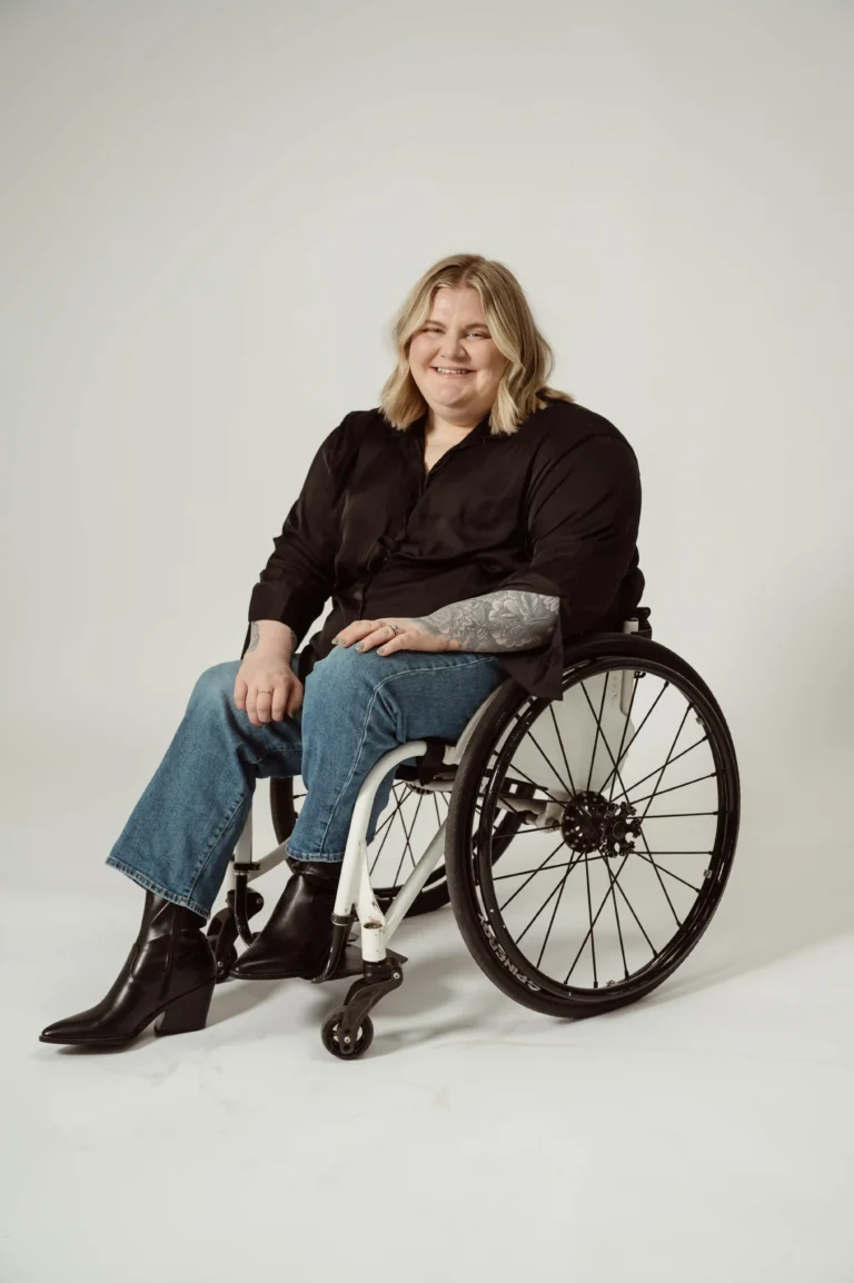 A person with blonde shoulder-length hair smiles while seated in a wheelchair. They are wearing a black long-sleeve blouse, blue jeans, and black ankle boots with a slight heel. Their left arm has visible tattoos, and they are resting their hands on their lap. The background is a neutral, light-colored backdrop.