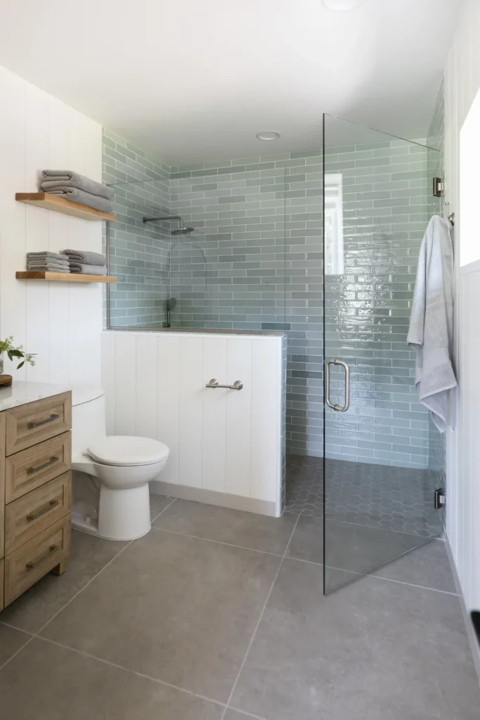Modern bathroom featuring a glass-enclosed shower with light green subway tiles on the walls and small hexagonal tiles on the shower floor. The bathroom has white vertical paneling on the lower walls, a natural wood vanity with drawers and silver handles, and open wooden shelves holding folded grey towels. The floor is covered in large gray tiles, and a towel hangs on a hook by the glass shower door.