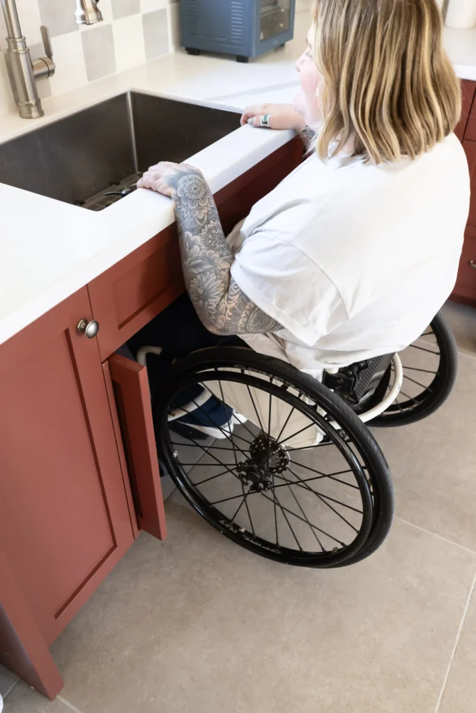 A person in a wheelchair is positioned at a kitchen sink, with red cabinets underneath that have an open space to allow for wheelchair access. They are wearing a white t-shirt and have detailed tattoos on their arms. The countertop is light-colored, and a faucet is positioned above the deep sink. The setting appears to be an accessible kitchen layout designed for wheelchair users.