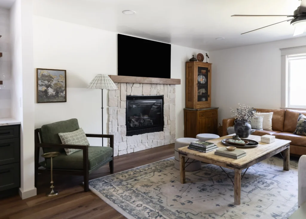 A cozy, rustic living room with a natural stone fireplace, topped by a wooden mantel and a black-framed TV. The space features a green armchair with a plaid pillow, a floor lamp with a patterned shade, and a leather sofa on the right. In the center, there is a large, wooden coffee table decorated with books, a vase with dried branches, and a ceramic bowl. A soft, patterned rug covers the hardwood floor. A wooden display cabinet stands next to the fireplace, adding to the room's warm and inviting ambiance.