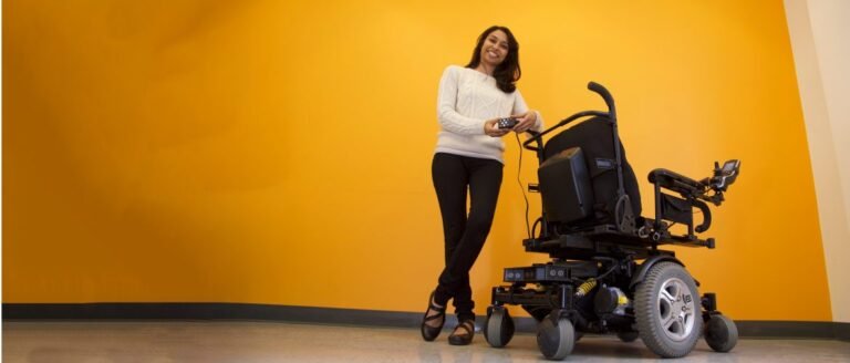 This image features a smiling woman standing next to a powered wheelchair. She is holding a controller, likely related to the wheelchair. The backdrop is a solid yellow or orange wall, giving a warm and vibrant atmosphere. The wheelchair appears to be equipped with some technology or sensors, possibly indicating it has advanced features, such as autonomous or assisted mobility. The woman seems to be demonstrating or presenting the wheelchair.
