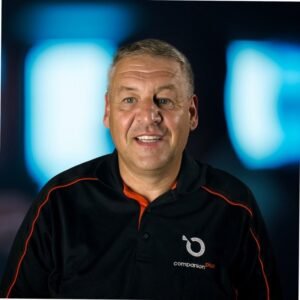 The image shows a smiling man, Eugene Cherny, with short, light brown or grayish hair. He is wearing a black polo shirt with orange details, and the logo "companion plus" is visible on the shirt's chest. The background appears to be out of focus, with blue and black gradient lighting, giving a soft, abstract effect. The man seems to be in a professional or corporate setting.