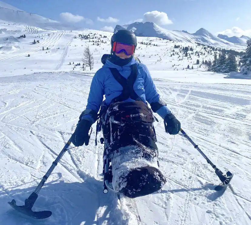 The image shows a person using adaptive ski equipment in a snowy, mountainous environment. They are seated in a sit-ski (mono-ski), a specialized piece of equipment used by individuals with limited lower body mobility for skiing. The person is holding outriggers (adaptive ski poles) for balance and control while skiing. They are dressed warmly in winter gear, including a blue jacket, black gloves, a helmet, and ski goggles. The snow-covered mountains and clear skies in the background highlight a scenic, alpine setting.