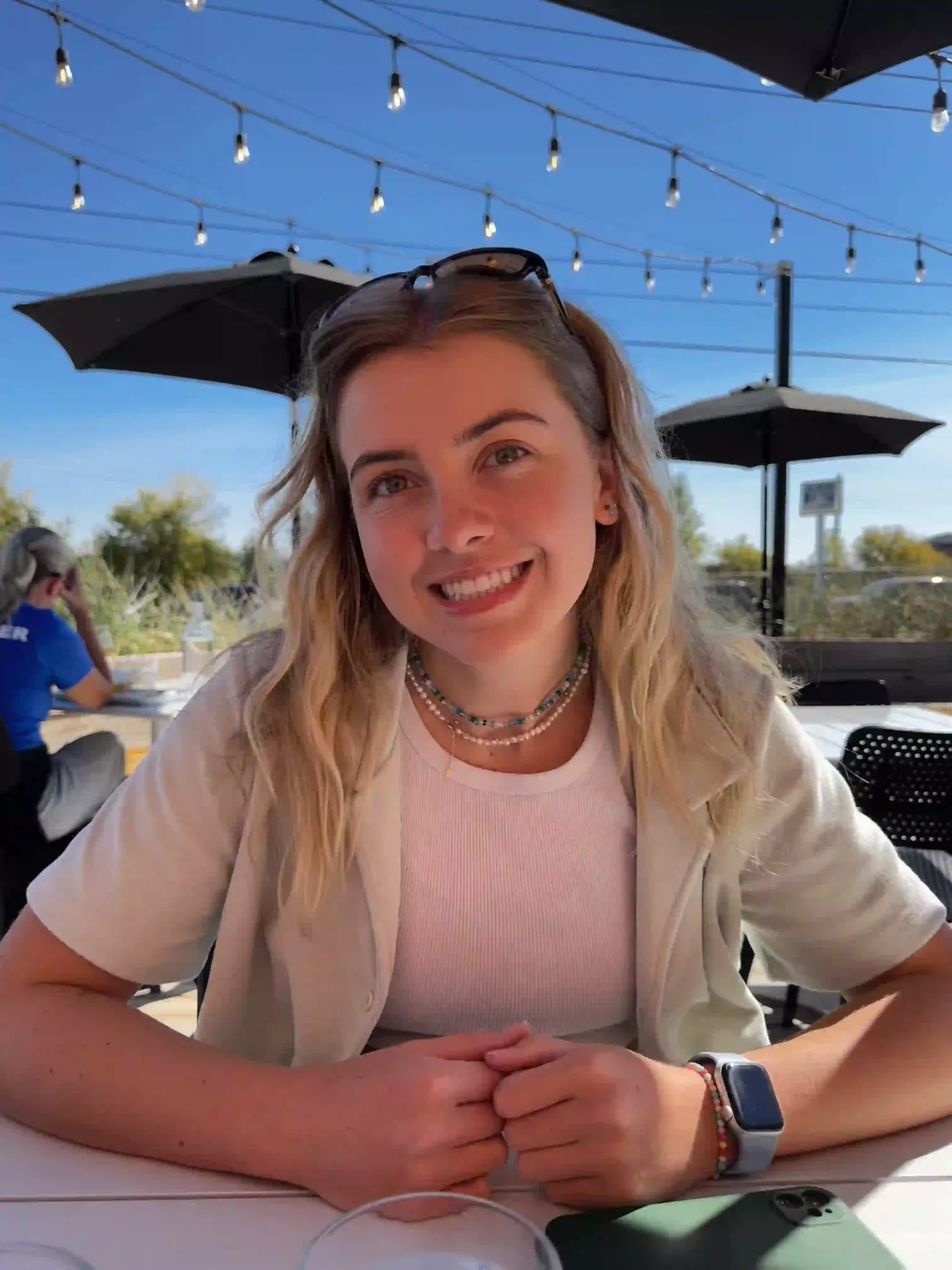 A young woman is sitting outdoors at a table under a clear blue sky with hanging string lights above. She has light blonde hair, styled in loose waves, and is wearing sunglasses on her head. She is smiling and dressed casually in a light-colored short-sleeved cardigan over a white top. She accessorizes with layered beaded necklaces, a watch on her wrist, and has her hands clasped together on the table in front of her. There are umbrellas and other diners visible in the background, creating a relaxed, friendly atmosphere.