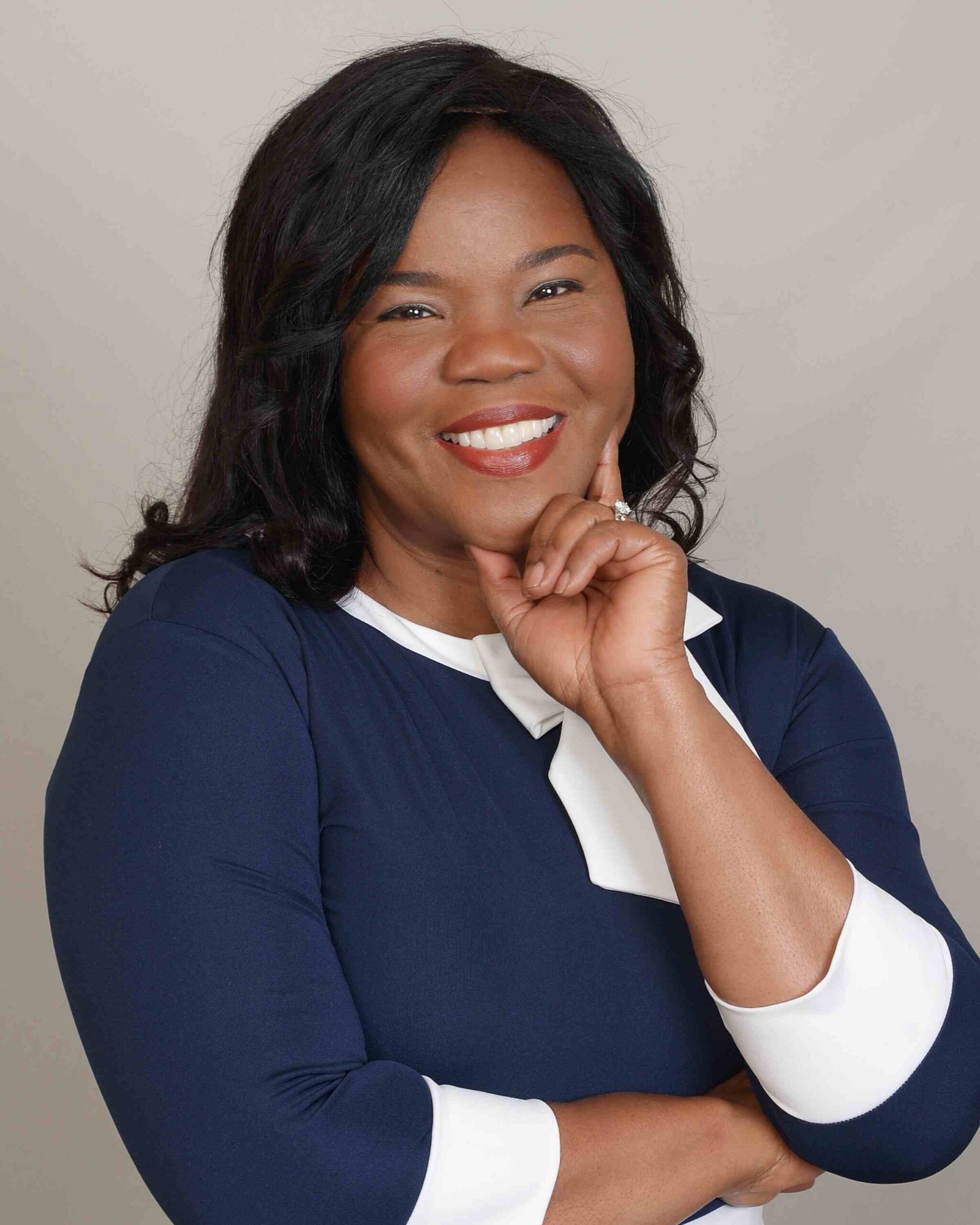The image shows a woman smiling confidently with her hand resting near her chin. She has medium-length black hair and is wearing a navy blue dress with white accents at the collar and cuffs. Her expression conveys warmth and professionalism, and she is posed against a neutral background.