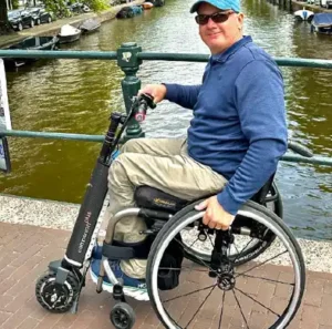 A man in a wheelchair is shown smiling while sitting on a bridge by a canal. He is wearing a blue long-sleeve shirt, light-colored pants, sunglasses, and a cap. His wheelchair is equipped with an electric assist attachment, giving it a scooter-like front wheel, allowing him to move more easily. The canal behind him has boats and is surrounded by greenery, suggesting a peaceful urban setting, possibly in a European city.