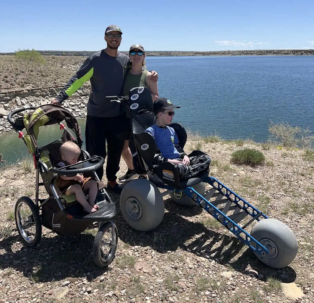 A family is outdoors near a lake on a clear, sunny day. Two adults are standing, one man and one woman, both wearing sunglasses and caps. The man has his arm around the woman. In front of them are two children in special strollers: one baby in a regular stroller on the left, and an older child in a larger off-road wheelchair with big balloon-like tires on the right. The scene shows a sense of adventure and togetherness, with a backdrop of blue water and a rocky shoreline.