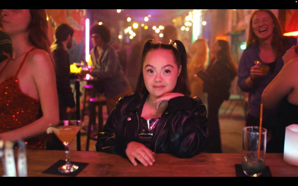 A young woman with Down syndrome is sitting confidently at a bar in a vibrant, lively setting. She is wearing a black leather jacket and has her hair styled in two high ponytails. The scene around her features colorful neon lights, blurred people talking and laughing in the background, and a lively bar atmosphere. A drink is in front of her on the bar, and she has a calm, composed expression, resting her chin on her hand as she looks directly at the camera. The scene conveys a sense of inclusion, confidence, and celebration.