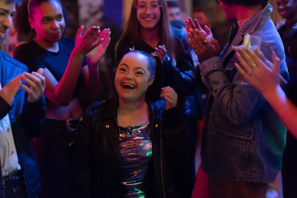 A group of friends is gathered at a party, with colorful lighting creating a lively atmosphere. In the center of the image, a young woman with Down syndrome is smiling joyfully, clearly enjoying the moment. She is dressed in a shiny, metallic outfit with a black leather jacket and has her hair styled in fun, playful pigtails. Surrounding her are other people clapping and cheering, all engaged in celebration. The mood is vibrant, full of energy, and inclusive, with everyone sharing in the happiness.
