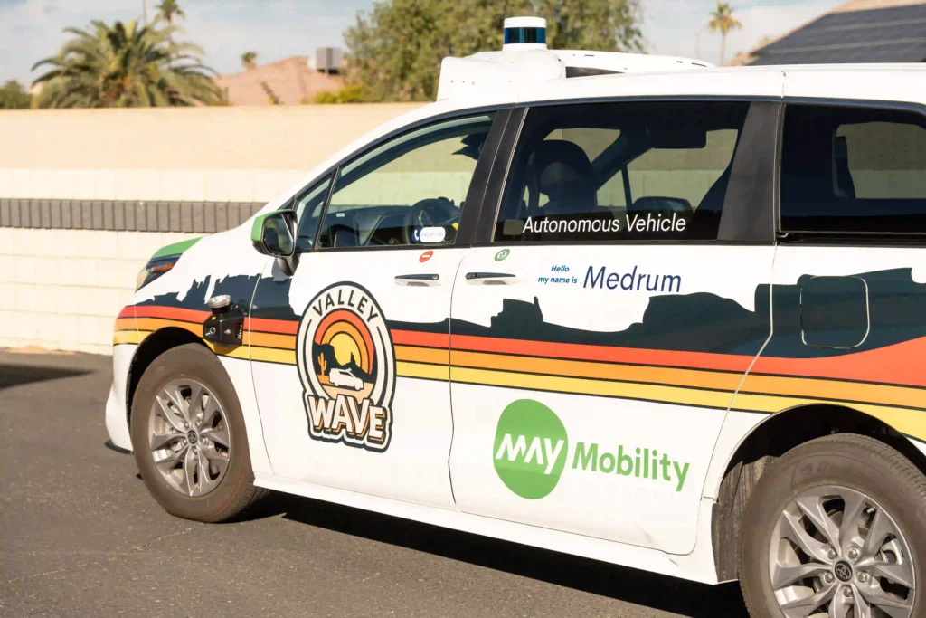 The image shows a white van marked with various logos and text, suggesting it is an autonomous vehicle. On the side of the vehicle, it reads "Autonomous Vehicle" in black text, and underneath it, there's text saying "Hello, my name is Medrum" in blue. The vehicle has the branding "Valley WAVE" with a logo featuring a sunset or sunrise over mountains and a reclining figure, and another logo "May Mobility" is visible in green text. The van appears to be outfitted with sensors, likely for autonomous driving. It is parked in an outdoor setting, with trees and houses visible in the background.