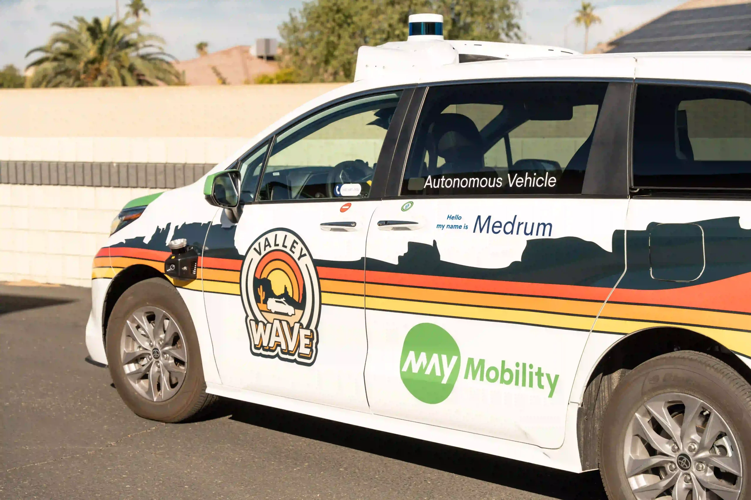The image shows a white van marked with various logos and text, suggesting it is an autonomous vehicle. On the side of the vehicle, it reads "Autonomous Vehicle" in black text, and underneath it, there's text saying "Hello, my name is Medrum" in blue. The vehicle has the branding "Valley WAVE" with a logo featuring a sunset or sunrise over mountains and a reclining figure, and another logo "May Mobility" is visible in green text. The van appears to be outfitted with sensors, likely for autonomous driving. It is parked in an outdoor setting, with trees and houses visible in the background.