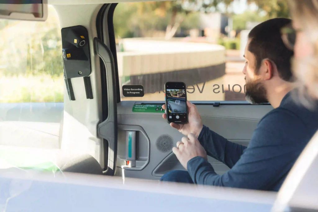A man with a beard is seated in the back of a vehicle, holding up a smartphone to take a photo or video through the window. A sign next to him reads "Video Recording In Progress." He is sitting near a small speaker or ventilation system built into the car's interior. The image captures a portion of a second person, partially visible in the foreground. The scene outside the vehicle shows a blurry view of a sunlit outdoor area. The interior of the vehicle also appears to have safety or monitoring equipment mounted to the side.