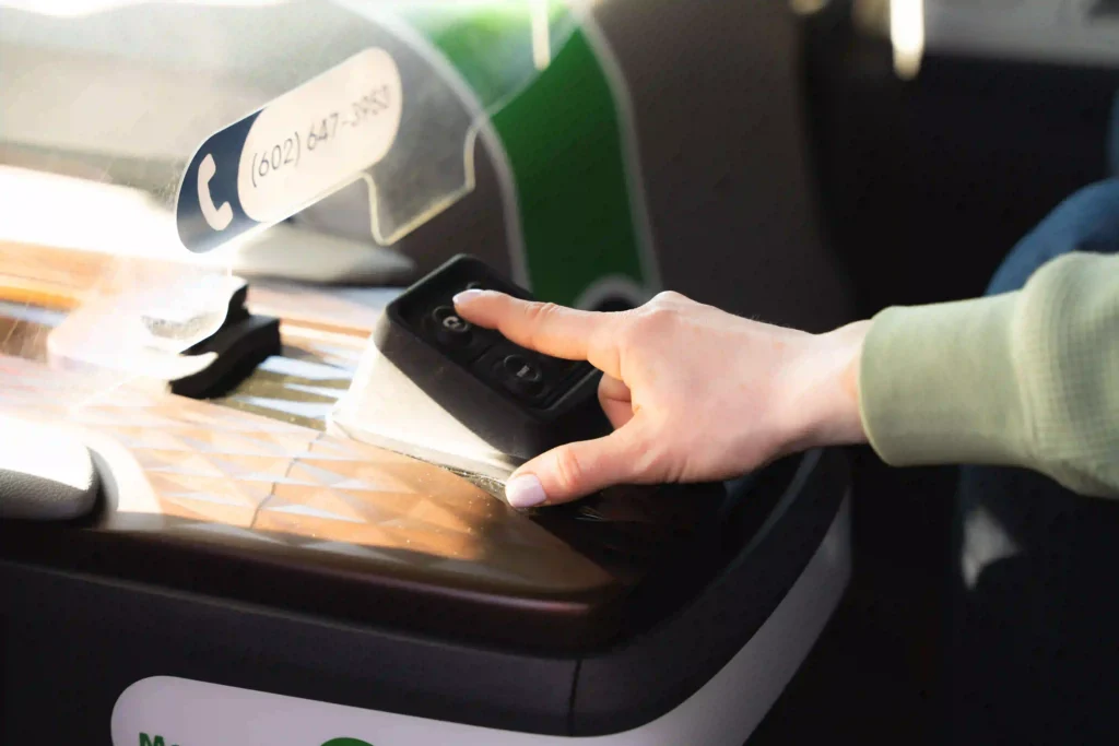 This image shows a close-up of a person's hand pressing a button on what appears to be a console or control panel. The hand is reaching for a rectangular remote control with visible buttons. A protective, transparent divider with a phone number on it is placed in the background. The surface under the hand has a textured, geometric pattern. The person is wearing a green sleeve.