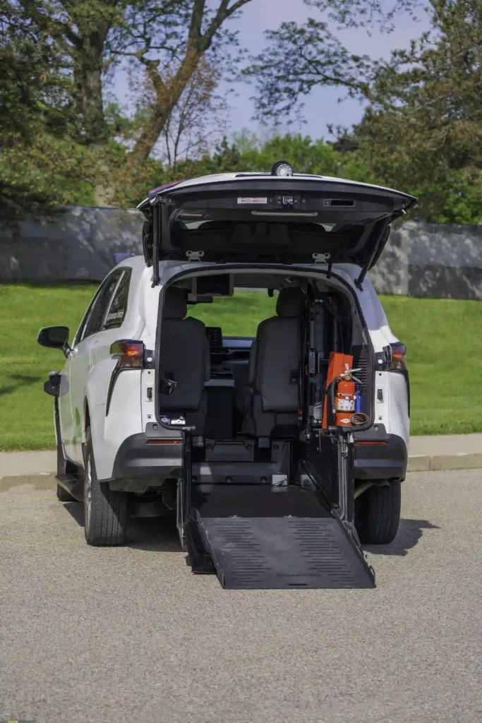 The image shows the rear of a white vehicle, likely a van or SUV, with the back door fully opened. A ramp extends from the rear to the ground, indicating the vehicle is likely modified for wheelchair accessibility. Inside the vehicle, two front seats are visible, and there is a fire extinguisher mounted on the side. The surrounding area is outdoors with trees and grass in the background. The image highlights the accessibility features of the vehicle.