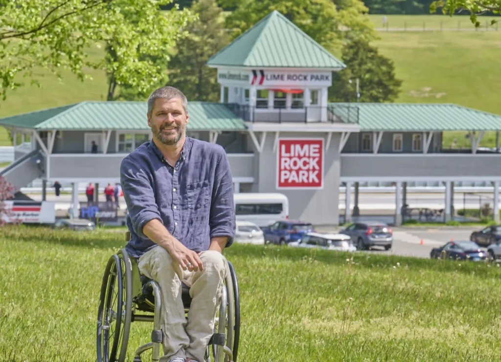 A man in a wheelchair is sitting on a grassy hill in front of a raceway building with "Lime Rock Park" signage prominently displayed. He has short, slightly graying hair and a beard, and is wearing a casual blue long-sleeved shirt and light-colored pants. The background shows a parking lot with several cars and part of a racetrack, with trees and greenery surrounding the scene. The atmosphere is bright and sunny, indicating a warm, clear day.
