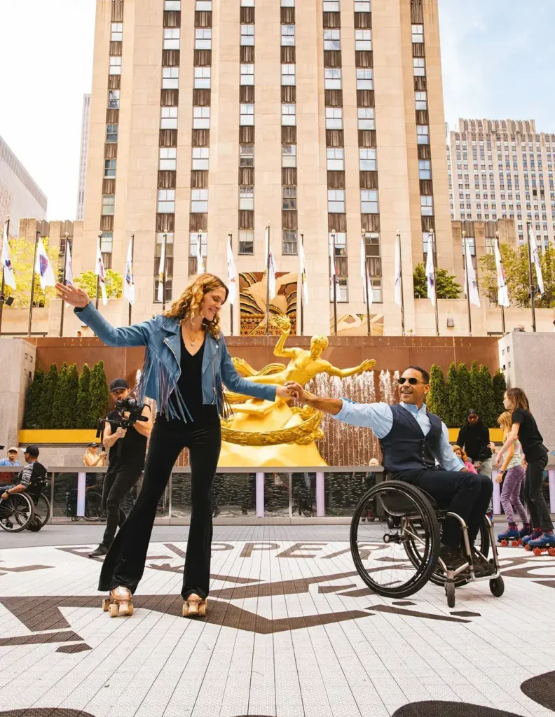 A man in a wheelchair and a woman roller skating are dancing together outdoors. The scene is set in front of the iconic golden statue of Prometheus at Rockefeller Center in New York City. The man is wearing a vest and sunglasses, while the woman is dressed in a blue fringed jacket and black pants. They are surrounded by other people, including a videographer, skaters, and spectators enjoying the atmosphere. The mood is lively and inclusive.