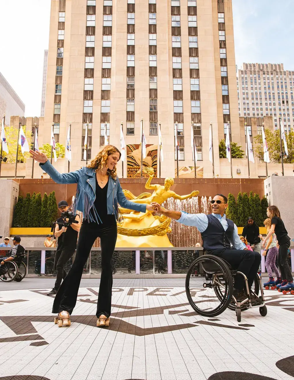 A man in a wheelchair and a woman roller skating are dancing together outdoors. The scene is set in front of the iconic golden statue of Prometheus at Rockefeller Center in New York City. The man is wearing a vest and sunglasses, while the woman is dressed in a blue fringed jacket and black pants. They are surrounded by other people, including a videographer, skaters, and spectators enjoying the atmosphere. The mood is lively and inclusive.