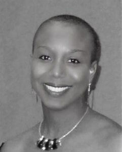 The image shows a black and white portrait of a smiling woman with short hair. She is wearing earrings and a necklace with large beads or ornaments. Her smile is warm, and she has an overall calm and pleasant expression.