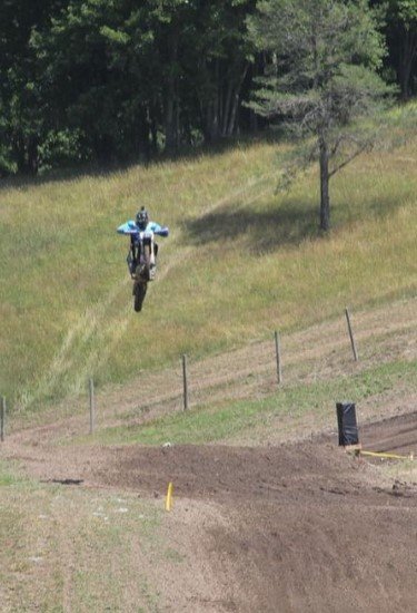 The image shows a person riding a dirt bike in mid-air, seemingly performing a jump on a motocross track. The rider is wearing a helmet and protective gear, and the surroundings feature grassy hills, a dirt track, and some fencing. The background has trees and open space, giving the impression that this is an outdoor motocross event.