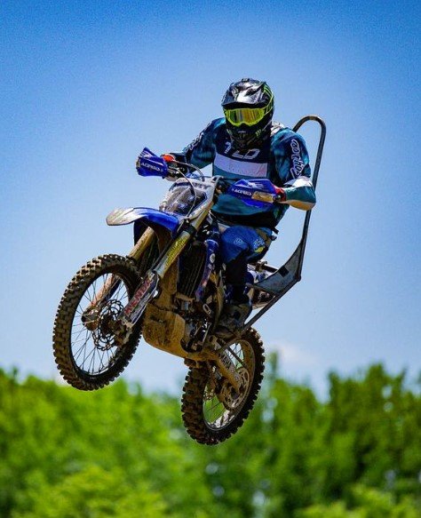 A motocross rider is captured mid-air, performing a jump on a dirt bike. The rider is wearing a blue and black motocross suit, a black helmet with a neon visor, and protective gear. The bike appears to be a Yamaha, with visible branding on the handguards. A roll cage or back brace seems to be attached to the rider's seat, likely for support or safety purposes. The background is a clear blue sky with a line of green trees below, highlighting the height of the jump. The image conveys a sense of speed, skill, and action.