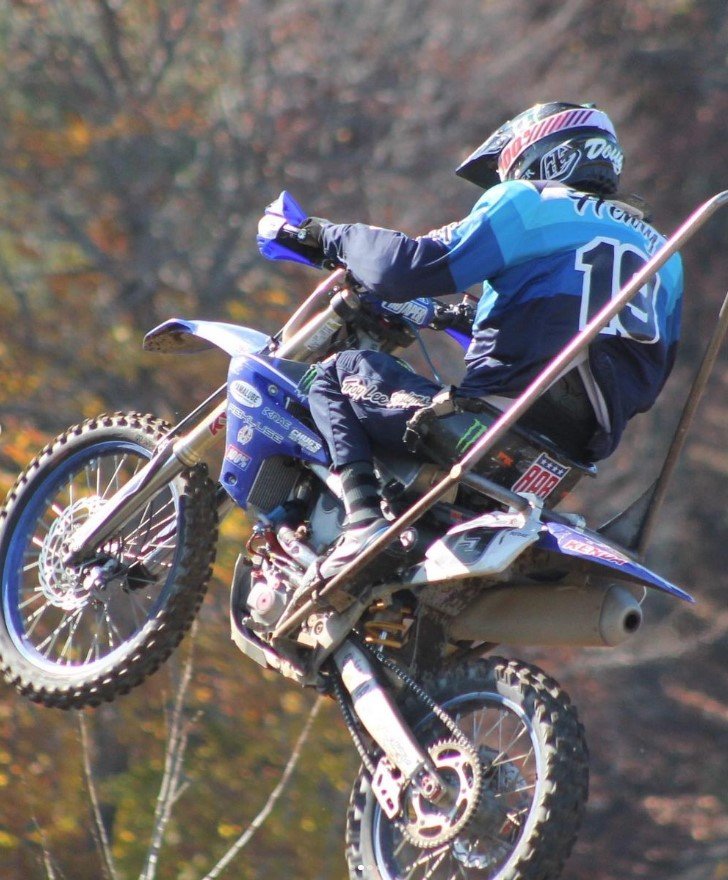 The image depicts a motocross rider in mid-jump, performing a wheelie on a dirt bike. The rider is wearing a helmet and a blue racing jersey with the number "19" visible on the back. The dirt bike is off the ground with both wheels lifted, showcasing the rider's skill and the bike's power. The surrounding background appears blurred, possibly due to the motion and the focus being on the rider. The scene takes place outdoors, likely during a motocross race or event.