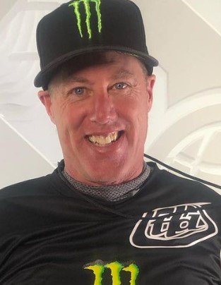 A man wearing a black cap with a Monster Energy logo and a matching black shirt is smiling. He has a friendly expression, and the background appears to have a light geometric design.