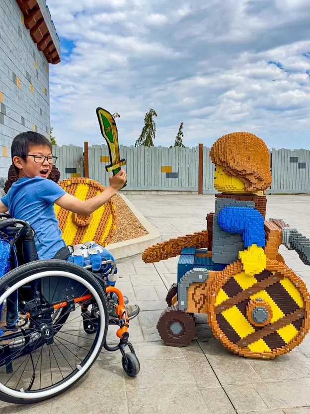 A young boy in a wheelchair enthusiastically holds up a toy sword while interacting with a life-sized LEGO figure. The LEGO figure is also in a wheelchair and holds a sword, creating a fun and inclusive scene. The setting is outdoors on a paved area with a fence and trees in the background under a partly cloudy sky. Both the boy and the LEGO figure appear to be playfully engaging in a mock battle.