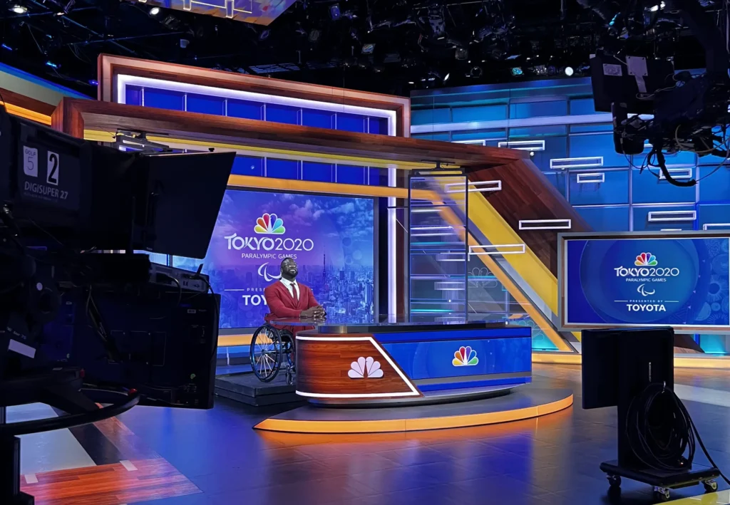 Broadcast studio set for the Tokyo 2020 Paralympic Games, featuring an NBC News desk with the Tokyo 2020 Paralympic logo displayed prominently on screens in the background. A host in a red suit and seated in a wheelchair is positioned at the desk, with professional studio lighting and cameras surrounding the set. The studio has a modern design with vibrant blue, yellow, and wood accents