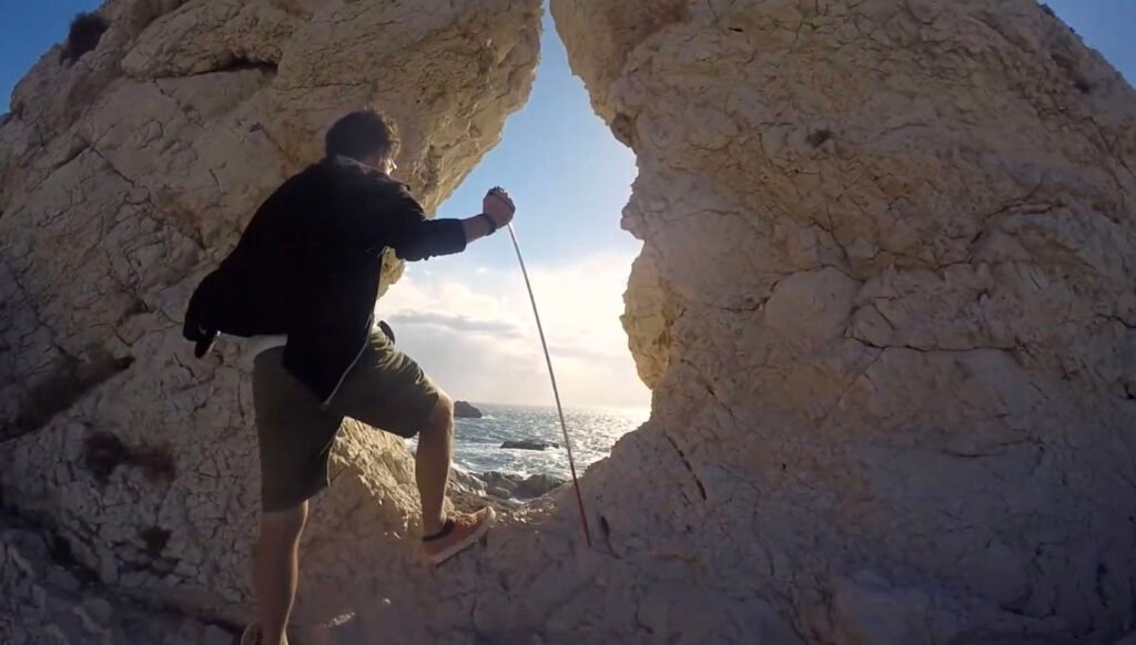 A person is climbing through a narrow rock crevice overlooking a bright and expansive view of the ocean. The individual is using a rope for support while stepping over rugged terrain. The surrounding rock formation is pale, and the sunlight creates a vibrant contrast with the sea in the distance. The scene evokes a sense of adventure and exploration.