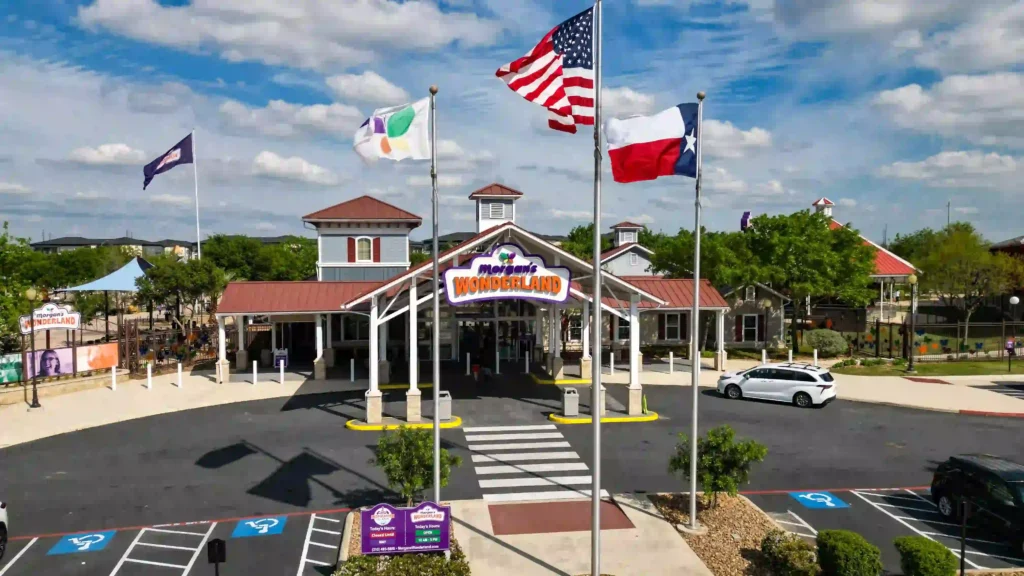 The image shows the front entrance of Morgan's Wonderland, an inclusive theme park. The main structure has a red-roofed building with a white-pillared entrance, and the park’s sign is prominently displayed above the entryway. Three flags are flying: the American flag, the Texas state flag, and another flag with colorful symbols. Below the flags, there is a car parked near designated handicapped parking spaces, indicating the park’s emphasis on accessibility. The setting is bright and sunny, with trees and playground equipment visible in the background.
