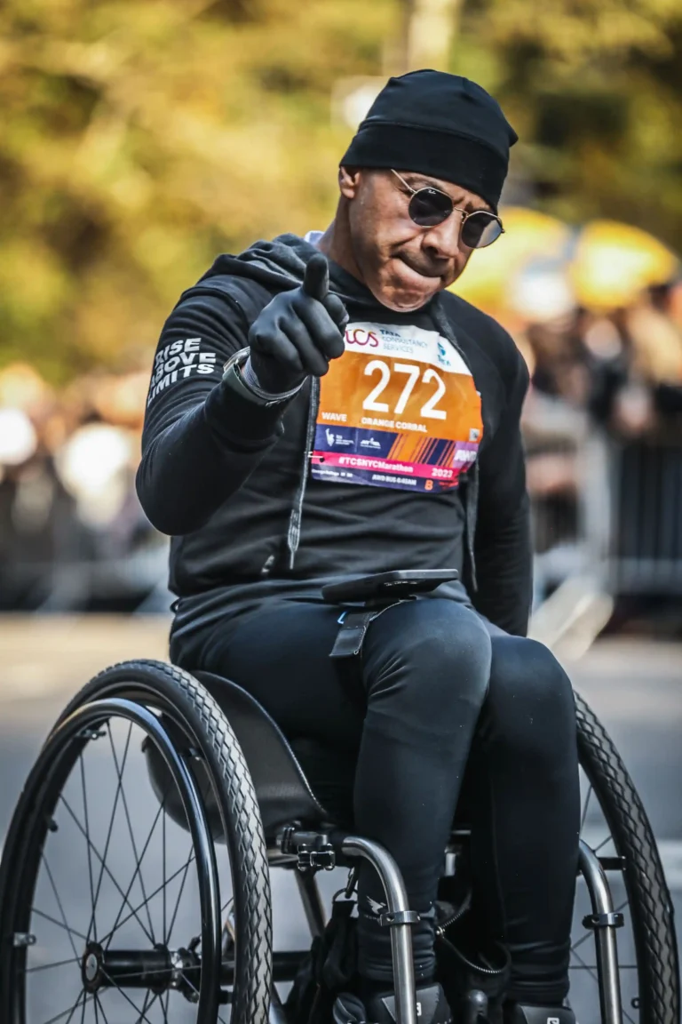 The image shows an athlete participating in a marathon, seated in a racing wheelchair. He is wearing black athletic gear, including a hat and gloves, and sunglasses. His race bib, numbered 272, is clearly visible and labeled with "TCS" and "New York City Marathon 2023." The athlete appears to be gesturing with his right hand, possibly giving a thumbs-up or pointing toward the camera. The words "Rise Above Limits" are written on his sleeve. The background shows a blurred crowd of spectators, indicating a lively event setting.