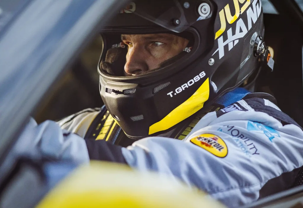 The image shows a race car driver, wearing a black helmet with a yellow accent and the name "T. GROSS" printed on the side of the helmet. The driver appears to be focused, looking intently ahead, likely seated inside a race car. The driver's suit features patches, including one for Pennzoil and another for "Mobility Innovation," as well as a harness across their chest. The image captures the intensity and concentration often associated with professional racing.