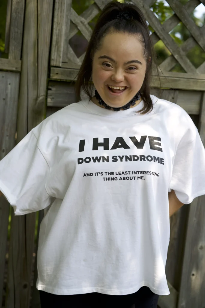 A young woman with Down syndrome is smiling brightly while wearing a white t-shirt with bold black text that reads, "I HAVE DOWN SYNDROME AND IT'S THE LEAST INTERESTING THING ABOUT ME." She has long dark hair pulled into a high ponytail and is standing outdoors in front of a wooden fence with latticework, creating a warm and positive atmosphere.